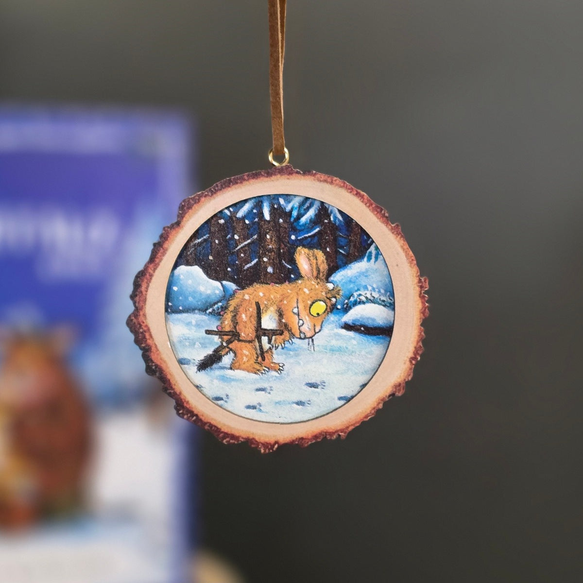 A circular Christmas ornament made from a natural wood slice with a hanging loop, featuring a painted winter scene of a brown Gruffalo in a snowy forest.