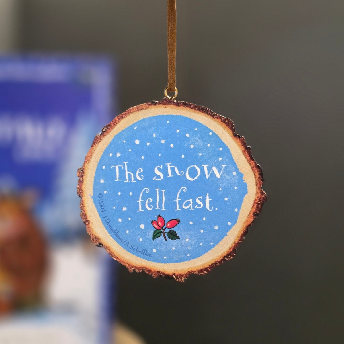 A round wooden Christmas ornament with a natural bark edge, featuring a blue painted center with white text reading 'The snow fell fast' and a small red and green berry illustration. A brown hanging loop is attached at the top, and the background is softly blurred.