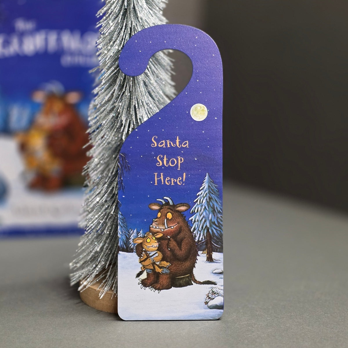 A printed Christmas door hanger featuring the Gruffalo and the The Little Nutkin in a snowy night scene with 'Santa Stop Here!' text, displayed next to a miniature silver Christmas tree.