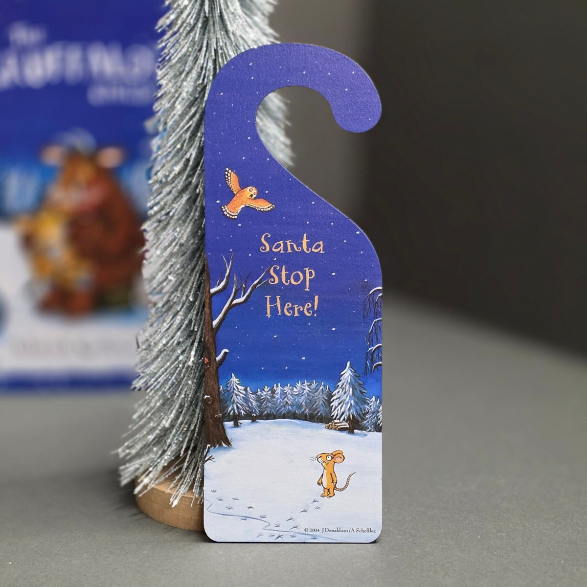 A Christmas-themed door hanger with a blue night sky, snowy landscape, and a small orange bird, printed with the text 'Santa Stop Here!'. It is shown standing upright next to a silver tinsel Christmas tree on a grey surface.