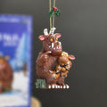 A Gruffalo-themed Christmas ornament featuring the brown Gruffalo holding a smaller Gruffalo baby, with a gold hanging string and holly decoration at the top, shown against a dark grey background with a blurred blue box in the side.
