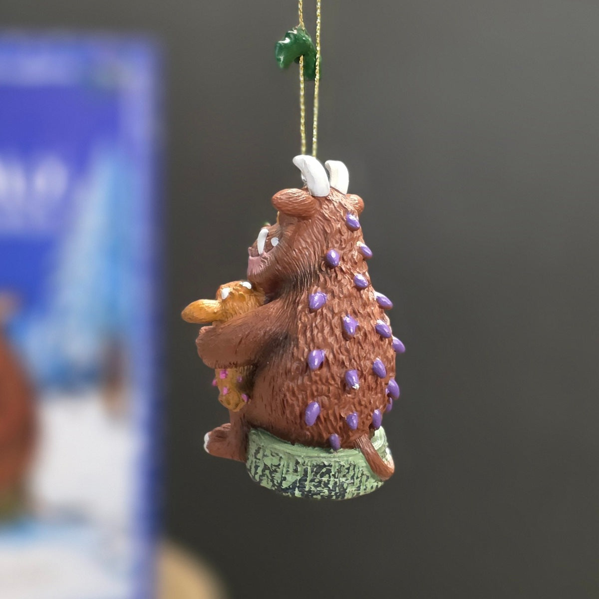 A small hanging ornament depicting a brown furry creature with purple spines, white horns, and a green base, suspended by a gold string against a dark blurred background.