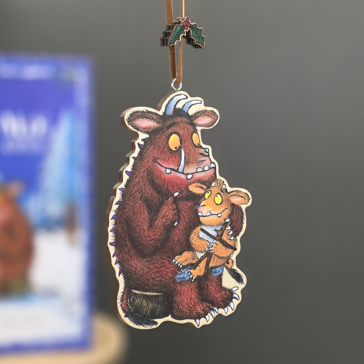 A die-cut wooden Christmas ornament featuring an illustration of the Gruffalo holding a small creature, hanging from a brown ribbon with a holly accent, against a blurred grey background.
