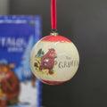 A spherical Christmas ornament featuring a cream-colored body with red top and bottom bands, decorated with an illustration of The Gruffalo and the title text. It hangs from a red ribbon against a dark grey background with a blurred The Gruffalo box in the background.