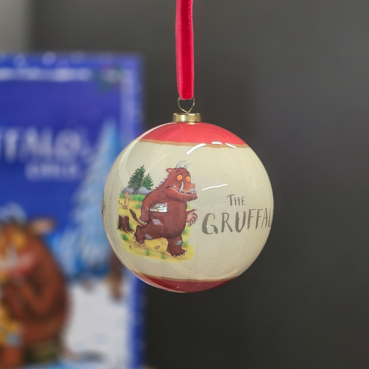 A spherical Christmas ornament featuring a cream-colored body with red top and bottom bands, decorated with an illustration of The Gruffalo and the title text. It hangs from a red ribbon against a dark grey background with a blurred The Gruffalo box in the background.