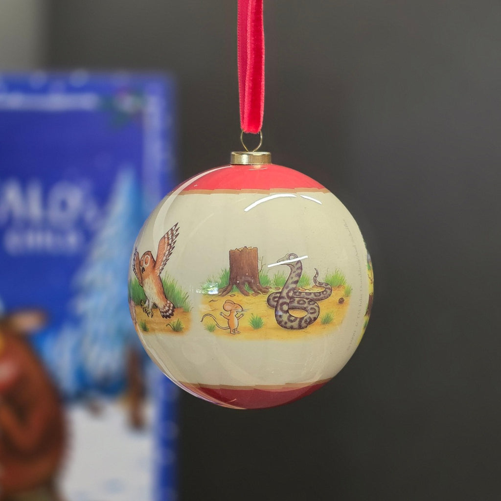 A round Christmas ornament with a red hanging ribbon, featuring a cream-colored background decorated with colorful illustrations of winter animals including a fox, a snake, a mouse, and a tree stump. A blue and white box is blurred in the background on the left.