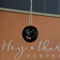 Black metal pumpkin-shaped ornament hanging against a orange background with text.