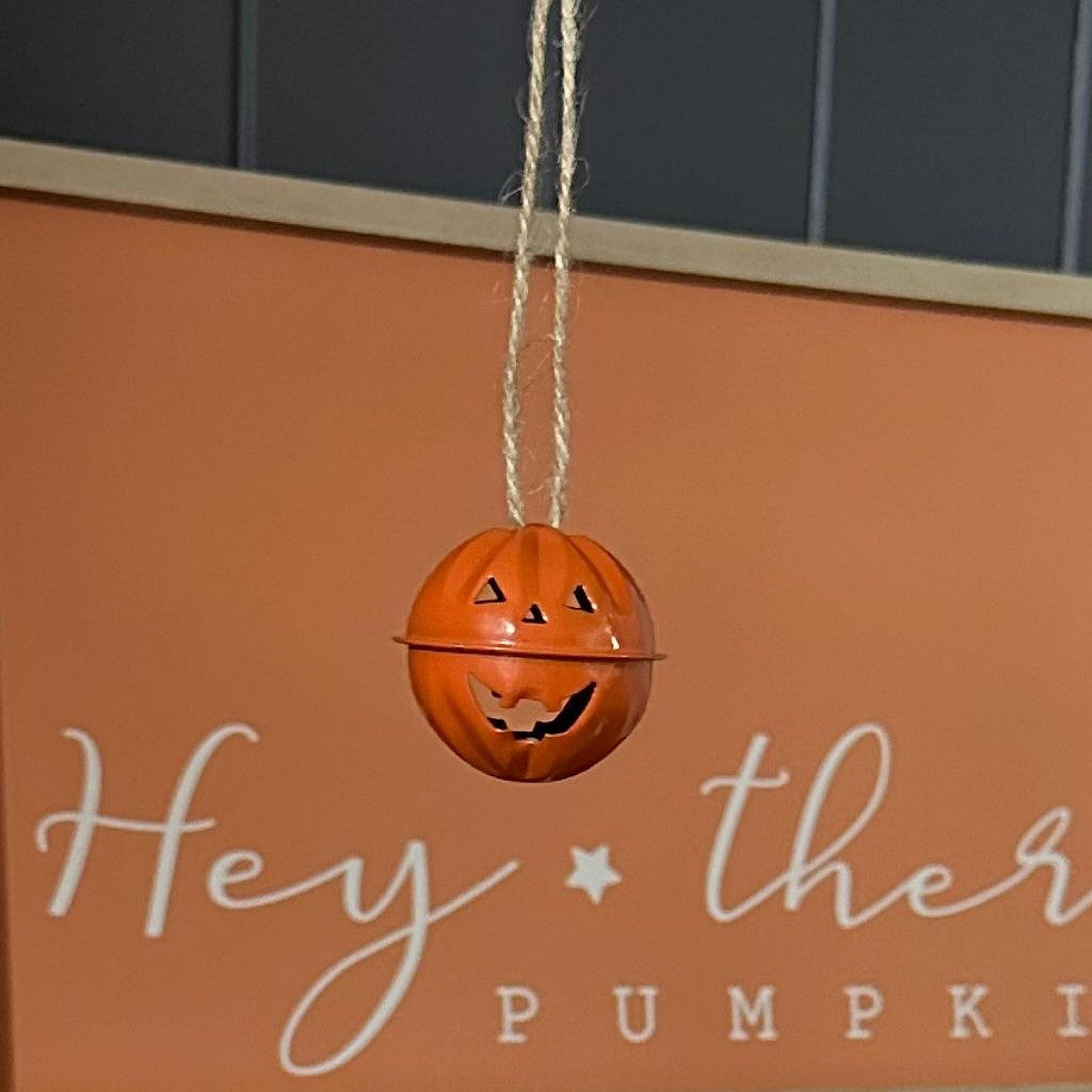 A small orange metal pumpkin-shaped hanging ornament with a carved jack-o'-lantern face, suspended by a twisted string in front of a warm orange sign with white script text.