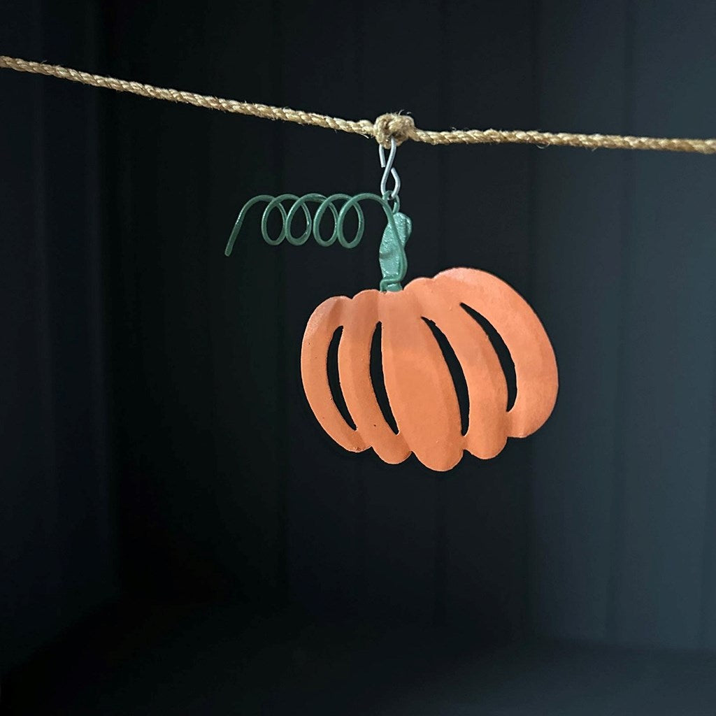 An orange pumpkin-shaped metal garland hanging with a green wire stem, suspended from a twine against a dark background.
