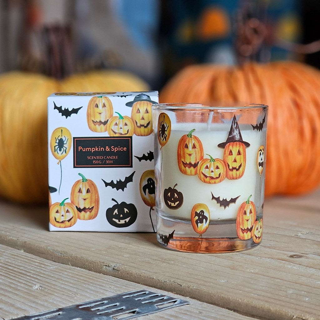 A Halloween-themed candle in a clear glass jar with a white wax base and orange jack-o'-lantern, spider, and witch illustrations, shown next to its printed packaging box on a wooden surface with pumpkins in the background.