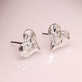A pair of silver heart-shaped stud earrings with a textured, hammered finish, displayed on a soft pink reflective surface.