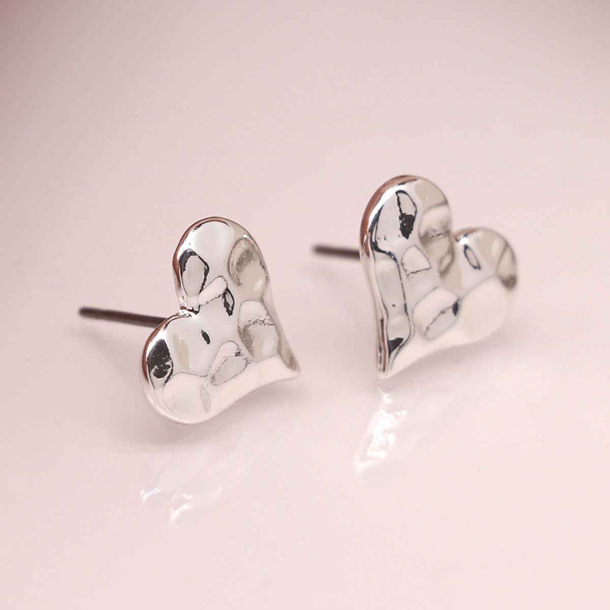 A pair of silver heart-shaped stud earrings with a textured, hammered finish, displayed on a soft pink reflective surface.