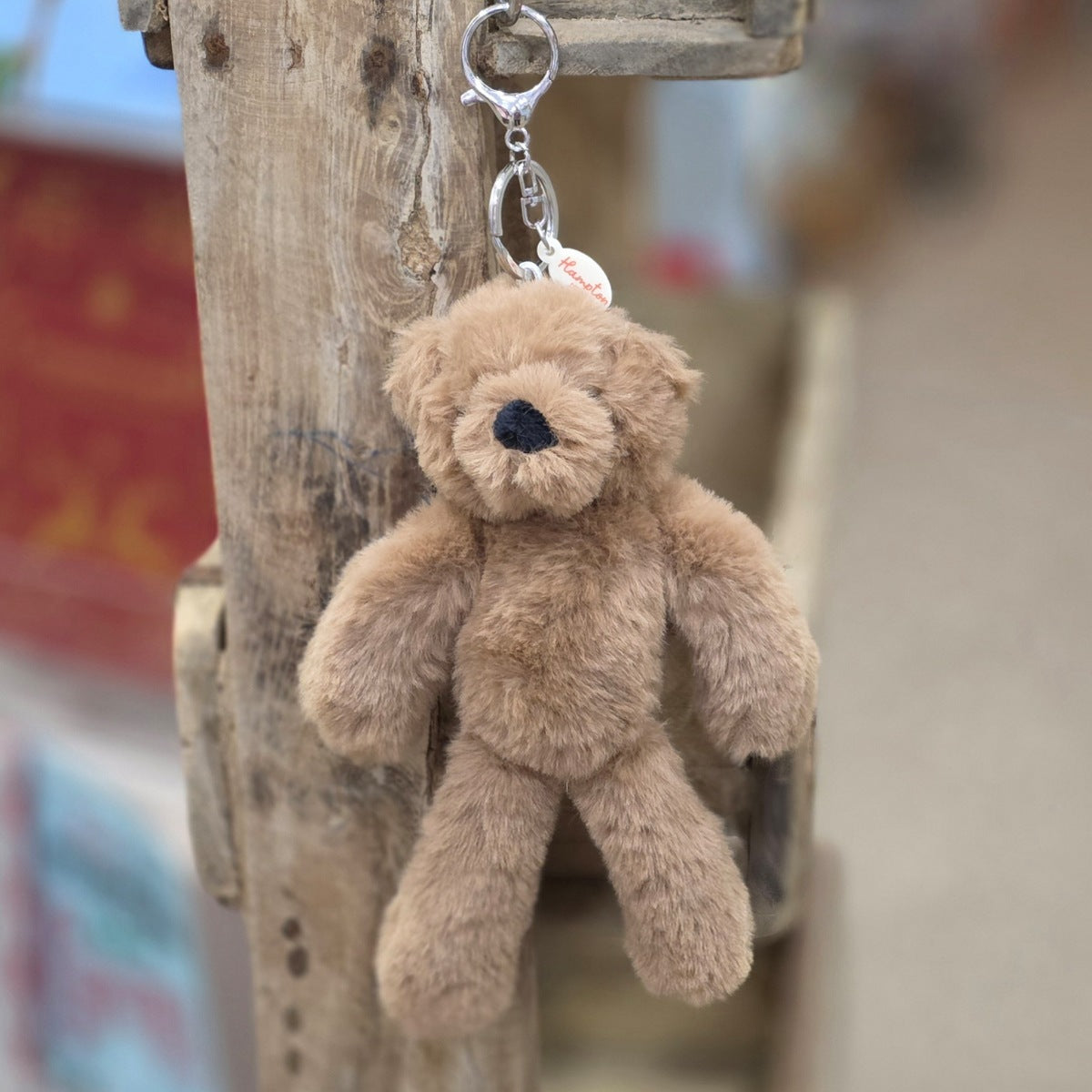 A brown plush teddy bear keychain hanging from a rustic wooden post. The keychain features a soft, fuzzy brown bear with black eyes and nose, attached to a silver metal key ring and a small round tag.

