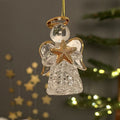 A clear glass angel Christmas ornament with gold-toned halo, wings, and star details, hanging against a softly blurred holiday background with warm lights.