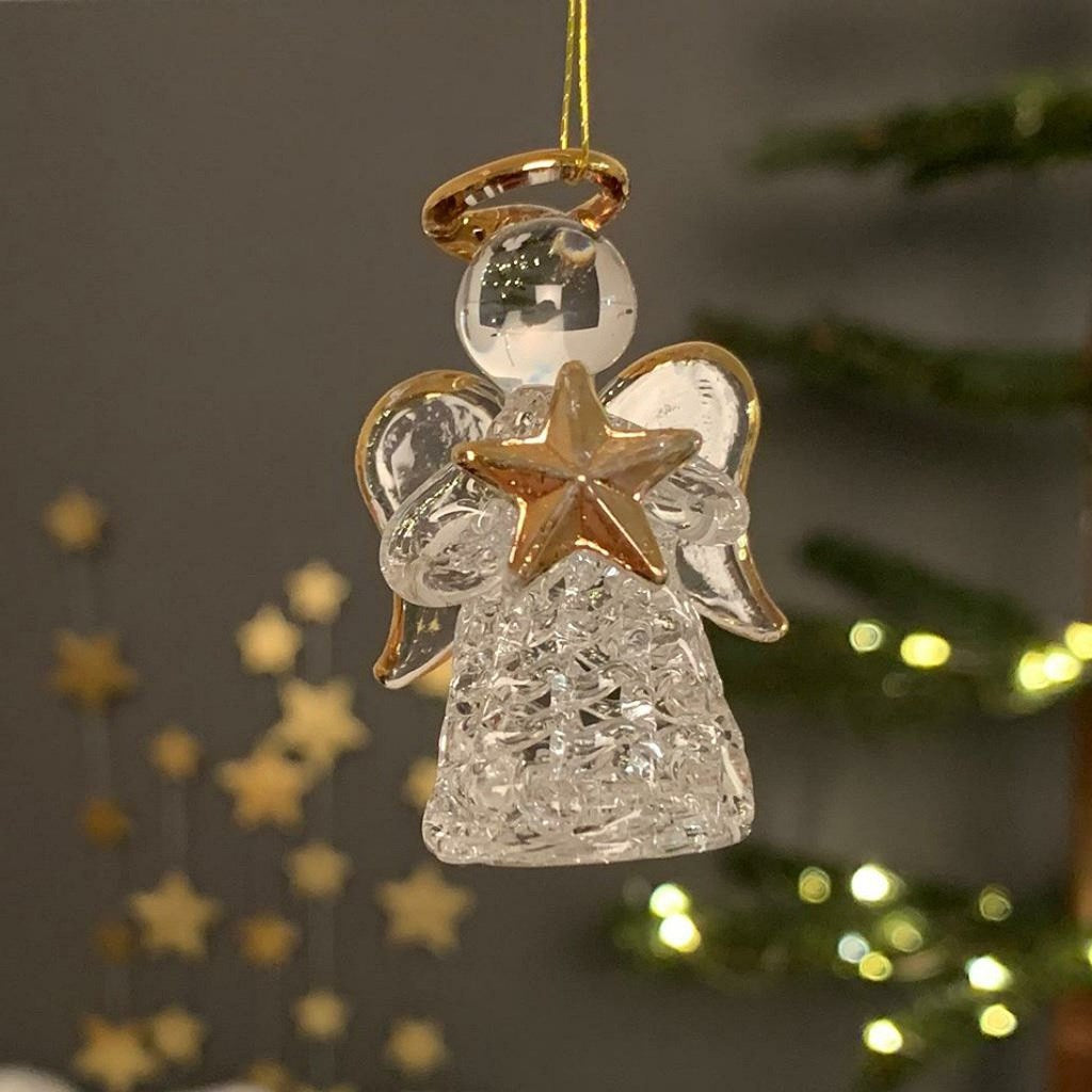 A clear glass angel Christmas ornament with gold-toned halo, wings, and star details, hanging against a softly blurred holiday background with warm lights.