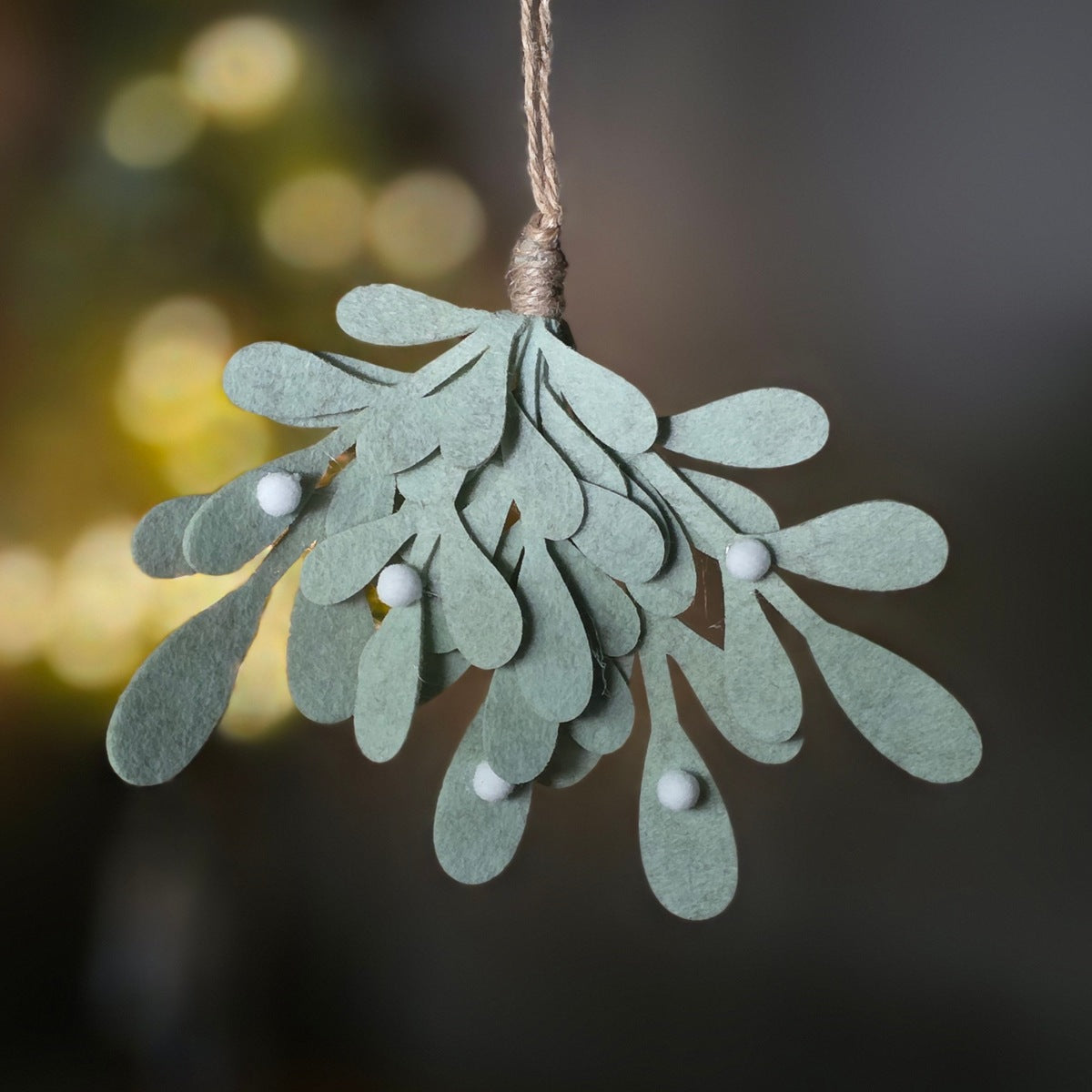 A felt mistletoe hanging ornament with small white berry details and a twine loop at the top, photographed against a softly blurred dark background with warm bokeh lights.