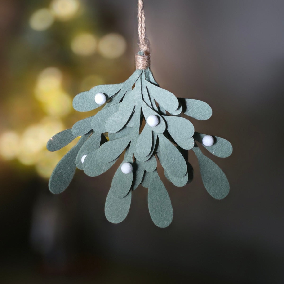 A mistletoe hanging decoration made from layered sage green felt leaves, adorned with small white pom-pom berries, hanging from a natural twine string against a dark, blurred holiday background.