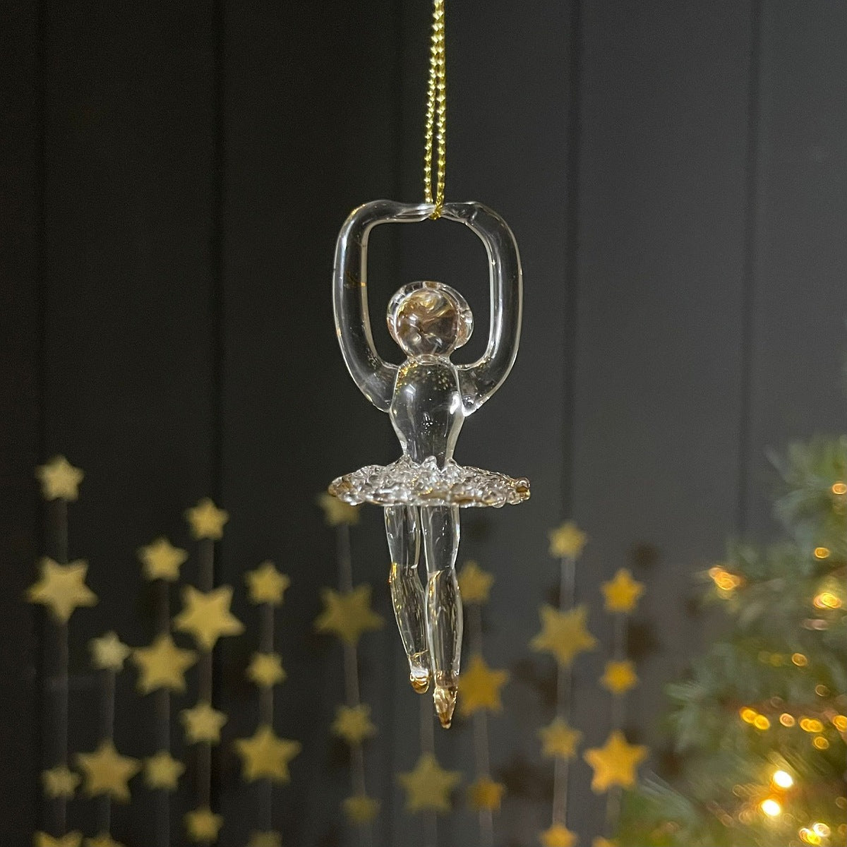 A clear glass ballerina Christmas ornament hanging from a gold chain against a dark paneled background with blurred gold star decorations and warm lights.