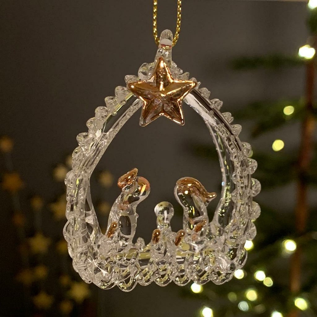 A clear glass Christmas nativity ornament with gold-toned star, animals, and figures, hanging by a gold string against a softly blurred holiday background.