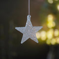 A silver glitter star-shaped hanging ornament suspended by a thin silver string, set against a dark, softly blurred background with warm yellow bokeh lights.