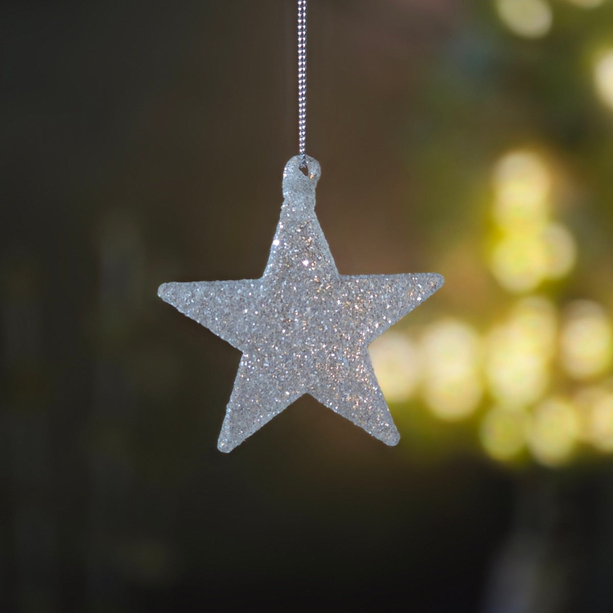 A silver glitter star-shaped hanging ornament suspended by a thin silver string, set against a dark, softly blurred background with warm yellow bokeh lights.