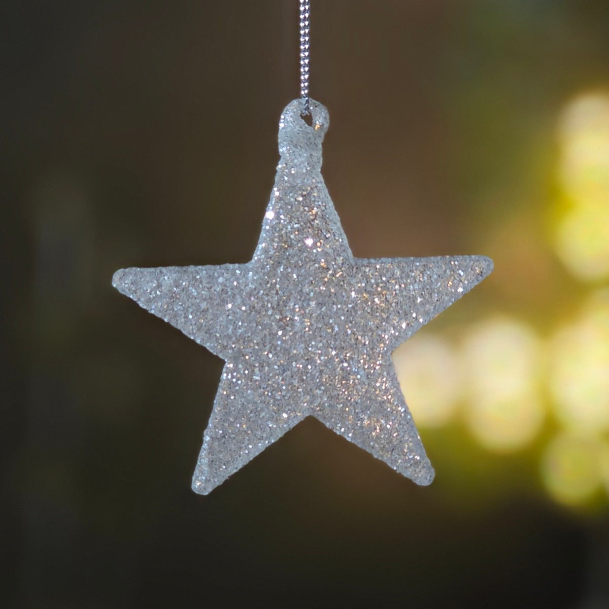 A glittery silver five-pointed star ornament hanging from a thin silver string against a dark, softly blurred background with warm yellow bokeh lights.