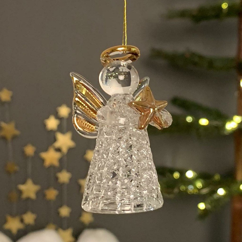 A hanging Christmas angel ornament made of clear textured glass with gold-toned wings, halo, and star detail, photographed against a softly blurred holiday background with lights and stars.