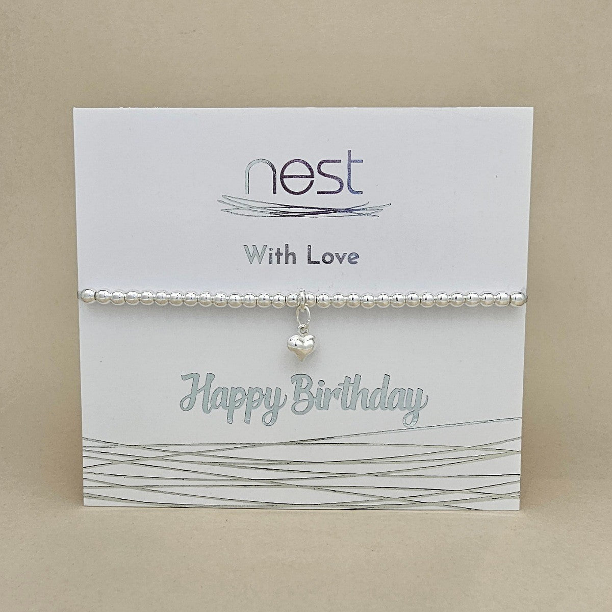 A silver beaded bracelet with a small heart charm, presented on a white card that reads 'nest With Love Happy Birthday' against a neutral beige background.