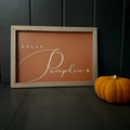 Wooden framed orange sign with 'Hello Pumpkin' text in white next to a small pumpkin on a dark surface.