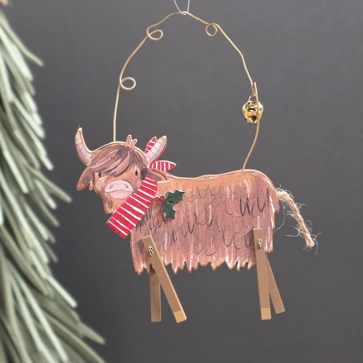 A wooden handcrafted Highland cow Christmas ornament hanging from a gold wire loop with a small gold bell. Decorated with a red and white striped scarf, a small green holly leaf, and wooden legs, set against a dark grey background with blurred pine needles on the left.