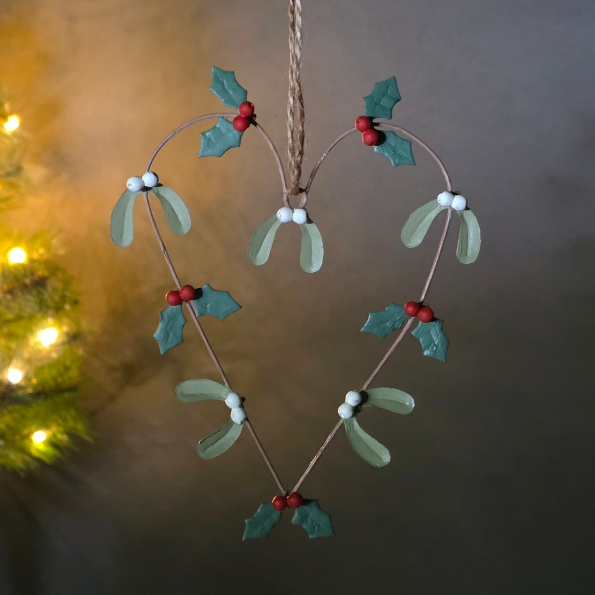 A heart-shaped Christmas hanging ornament made of thin brown wire, decorated with green mistletoe leaves, small red berries, and white bead accents, suspended by a twine string against a softly blurred background.