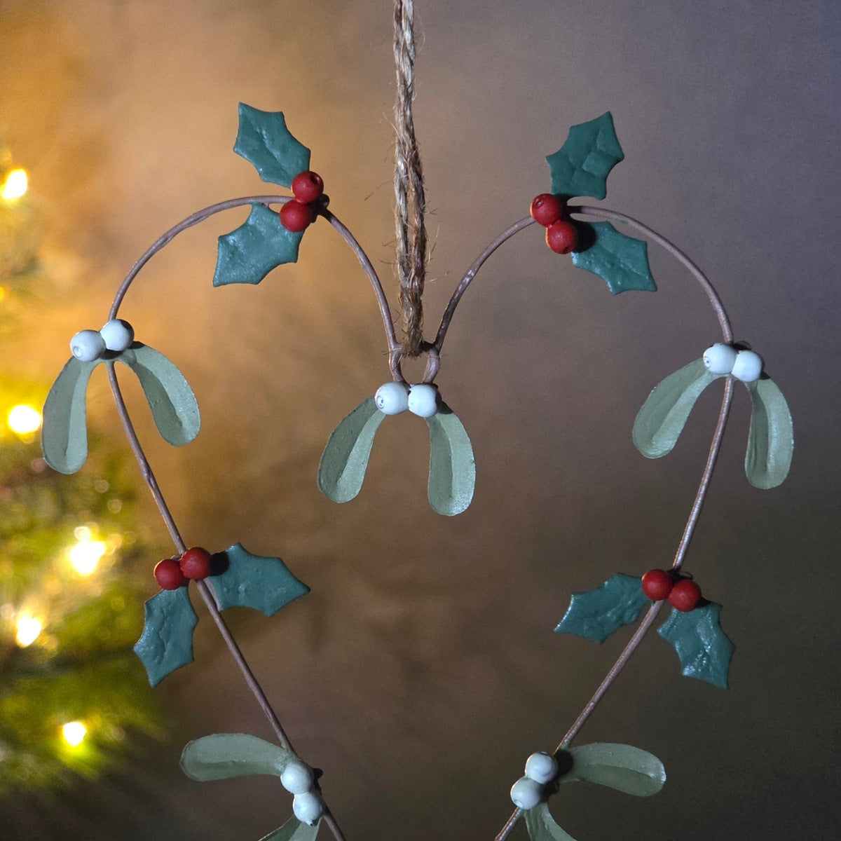 A heart-shaped Christmas hanging ornament made of thin brown wire, decorated with green mistletoe leaves, small red berries, and white bead accents, suspended by a twine string against a softly blurred background.