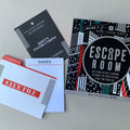 Escape Room kit contents arranged on a light gray surface, including a black instruction booklet, a white notes card, a white clue file with red accents, and a black-and-white Escape Room box with red and teal graphic accents.