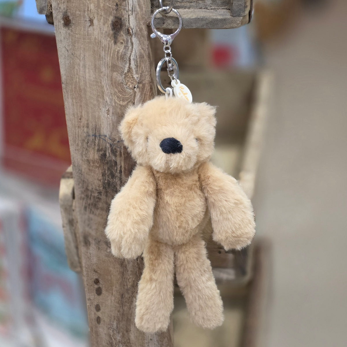 A light brown plush teddy bear keychain hanging from a rustic wooden post. The keychain features a soft, fuzzy light brown bear with black eyes and nose, attached to a silver metal key ring and a small round tag.
