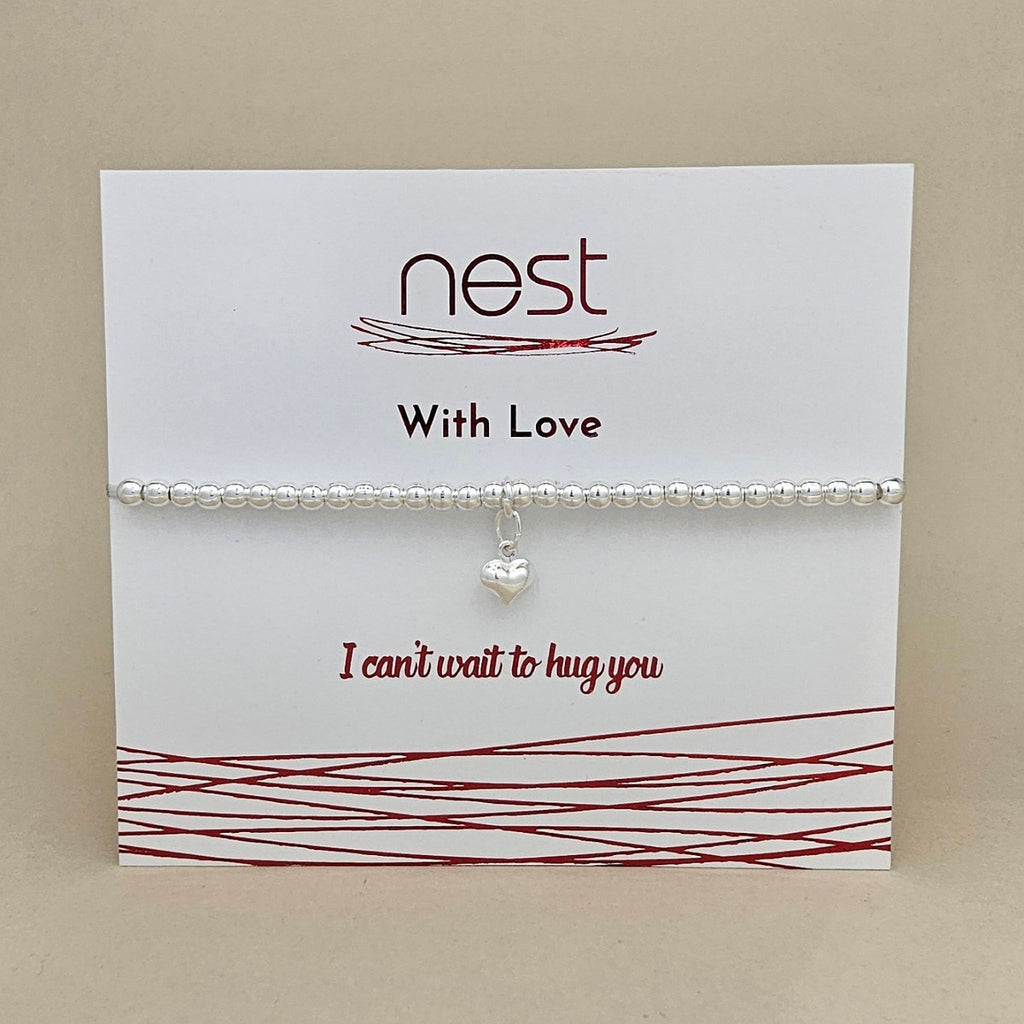 A silver beaded bracelet with a small silver heart charm, displayed on a white card from the brand 'nest' with red text and decorative lines, set against a neutral beige background.