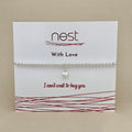 A silver beaded bracelet with a small silver heart charm, displayed on a white card from the brand 'nest' with red text and decorative lines, set against a neutral beige background.