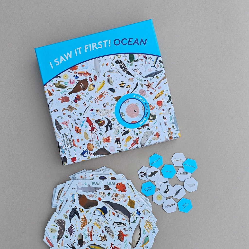 An educational children's game titled 'I SAW IT FIRST! OCEAN' shown on a neutral grey background, alongside a stack of illustrated ocean-themed tabs and small blue hexagonal cards.