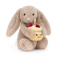 A plush toy bunny in warm taupe holding a small yellow cupcake character with a smiling face, red cherries, and a striped candle, photographed on a clean white background.