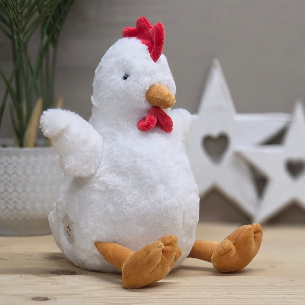 A white plush chicken toy with a red comb and wattle, orange beak and feet, sitting on a light wooden surface with a blurred indoor background featuring a plant and white star decor.