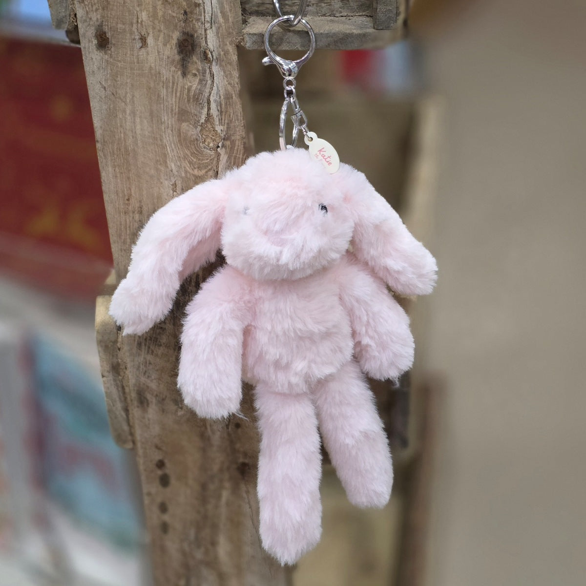 A small plush bunny keychain in light pink, hanging from a metal ring and clip on a rustic wooden post. The bunny has long floppy ears, a soft fuzzy texture, and a small attached tag.