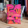 The book 'The Magic of Mushrooms' by Sandra Lawrence is standing upright on a wooden surface with a blurred background of books and household items. The cover features vibrant, stylized illustrations of pink and orange mushrooms on a black background with yellow text.