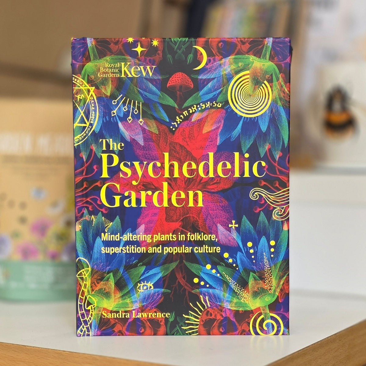 The Psychedelic Garden book by Sandra Lawrence, featuring a vibrant, multicolored psychedelic cover with yellow title text, displayed on a wooden surface with a blurred background.