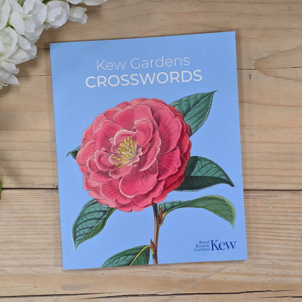 A book cover for 'Kew Gardens Crosswords' lies on a light wood surface, featuring a large pink flower with green leaves on a light blue background. White flowers are partially visible in the top-left corner.