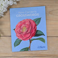A book cover for 'Kew Gardens Crosswords' lies on a light wood surface, featuring a large pink flower with green leaves on a light blue background. White flowers are partially visible in the top-left corner.