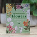 The Kew Gardens Flowers Colouring Book standing upright on a light wooden surface, with a blurred garden background featuring green leaves and potted plants.