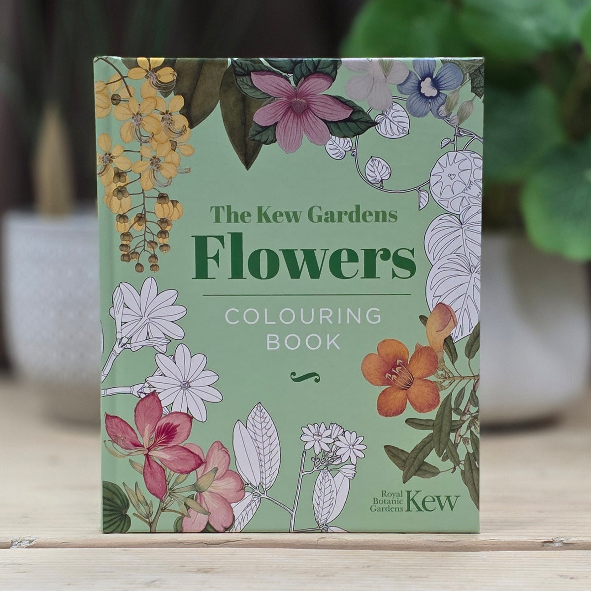 The Kew Gardens Flowers Colouring Book standing upright on a light wooden surface, with a blurred garden background featuring green leaves and potted plants.