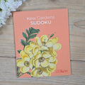 A book cover titled "Kew Gardens SUDOKU" lies on a light wood surface. The cover is peach-colored with a large illustration of yellow flowers and green leaves, and the text "Royal Botanic Gardens Kew" appears in the lower right corner.