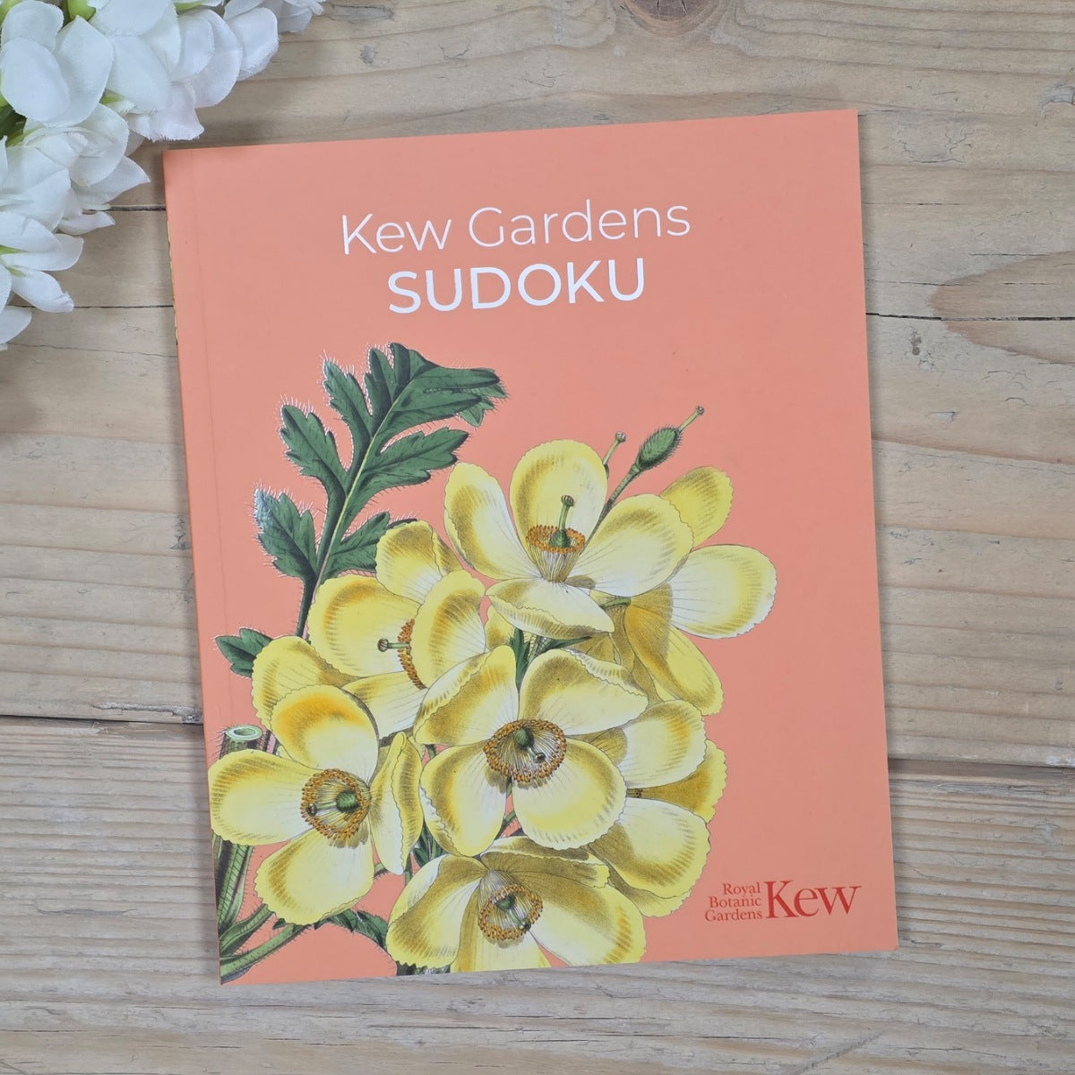 A book cover titled "Kew Gardens SUDOKU" lies on a light wood surface. The cover is peach-colored with a large illustration of yellow flowers and green leaves, and the text "Royal Botanic Gardens Kew" appears in the lower right corner.