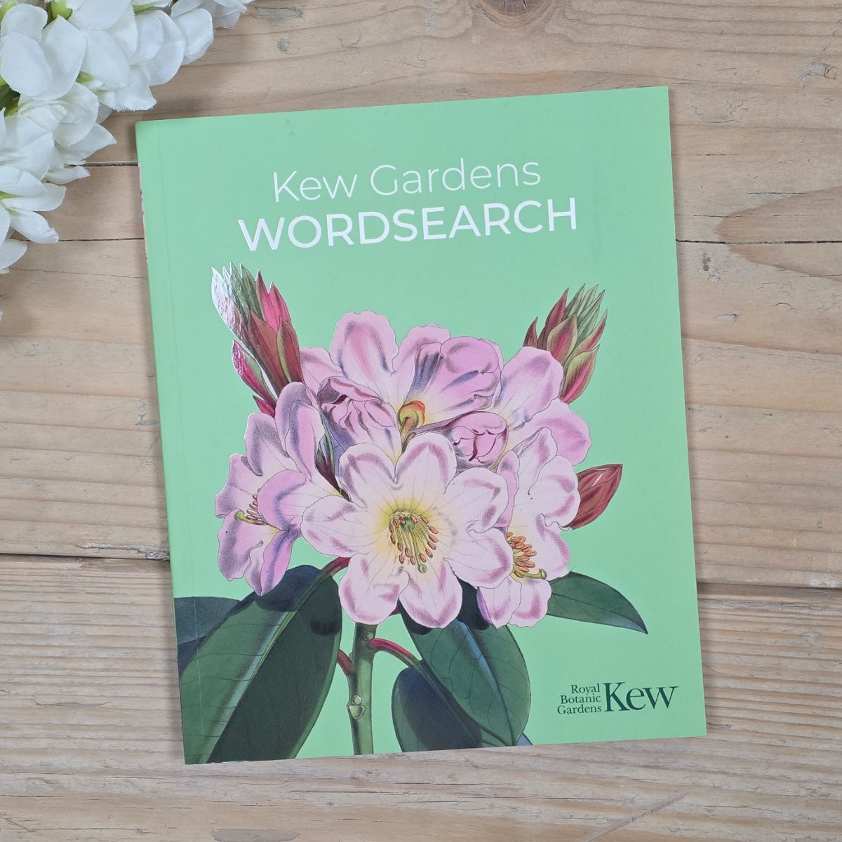 A Kew Gardens Wordsearch book or booklet lying on a light wood surface, with a mint green cover featuring a large pink flower illustration and white title text. White flowers are partially visible in the top-left corner.