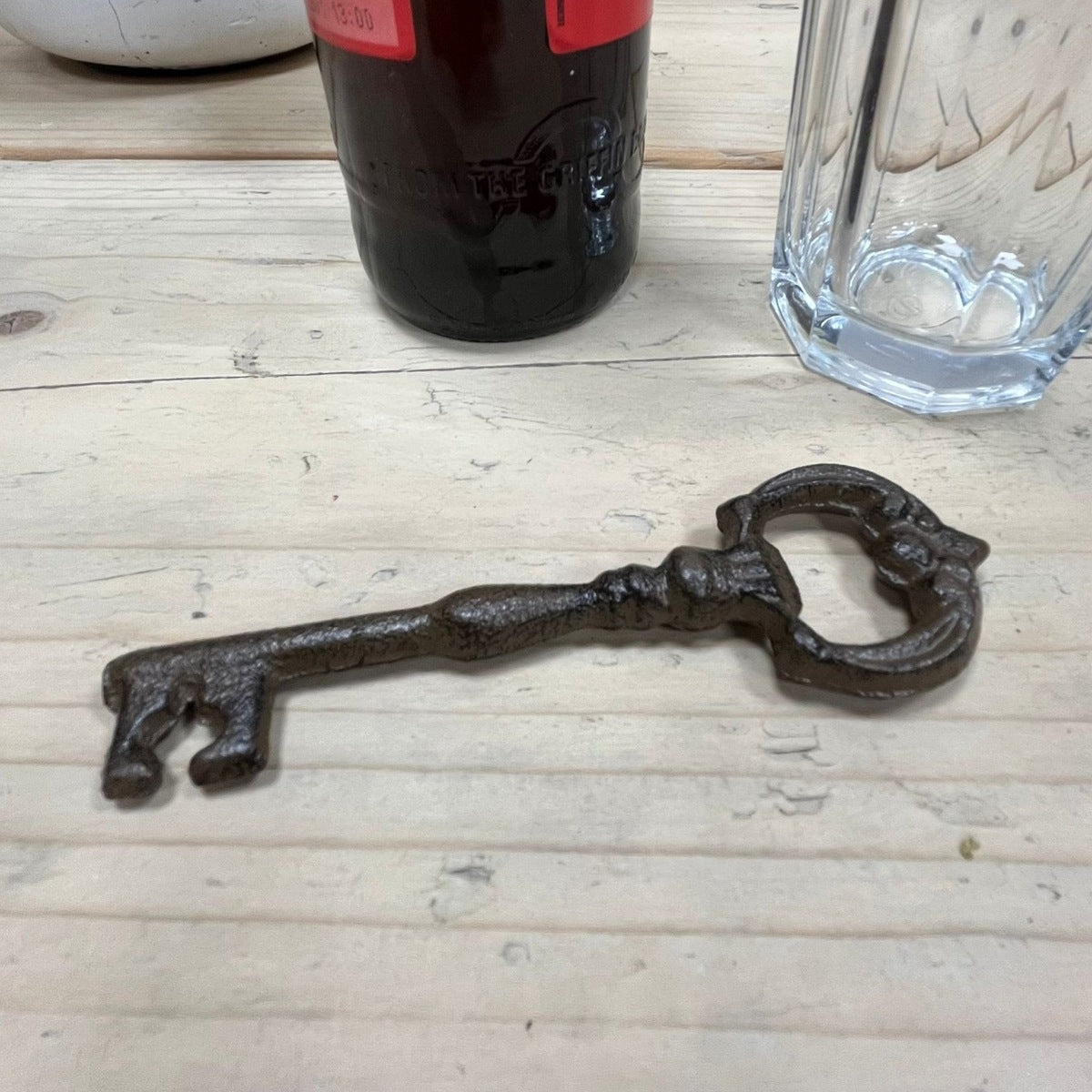 Decorative metal key-shaped bottle opener on a wooden surface with a bottle and glass in the background.